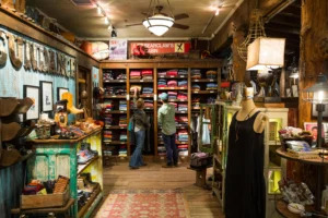 Inside a rustic, cozy shop with wooden interiors, featuring items like clothes, shoes, and accessories. banners and vintage décor enhance the nostalgic, cabin-like atmosphere. two customers are browsing.