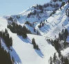 A scenic winter landscape showing snow-covered mountains with ski tracks and snow-covered pines under a blue sky. skiers are visible on the slopes, and a ski lift runs along the side.