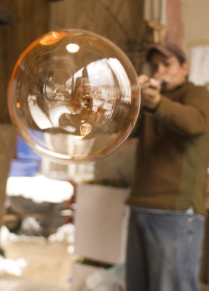 A glassblower focuses intently on shaping a large, nearly spherical glass bubble using a blowpipe, with workshop details softly blurred in the background.