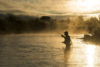 A silhouette of a fisherman, casting a line in a misty river during sunrise at Sundance Resort, with dense foliage and a glowing sky in the background.