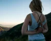 A woman practices yoga on a mountain, facing away from the camera. she holds her hands in a prayer position behind her back, overlooking a serene landscape at sunset.