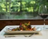 A romantic gourmet meal on a rectangular plate, accompanied by a glass of red wine, set on a wooden table with a blurred greenery background.