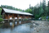 A serene wooden cabin with a large roof extends over a clear stream bordered by lush forests. white deck chairs are visible on the cabin’s porch, enhancing the peaceful nature retreat vibe.