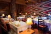 A cozy restaurant interior with a rustic cabin feel, featuring wooden beams, a large geometric-patterned rug on the wall, and multiple tables set with white linens and lit lamps.