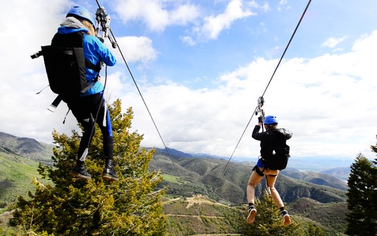 Two people zip-lining over a lush mountainous landscape, dressed in blue and black adventure gear with helmets, one person slightly ahead of the other.