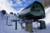 A ski lift station with an enclosed green lift cabin and an adjacent open chairlift carrying skiers, set against a snowy mountain landscape and a clear blue sky.