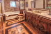 A vintage-style bathroom features a wooden vanity with a sink, ornate mirror, stained glass window, and a clawfoot bathtub. The floor showcases intricate wood designs, and a brick accent wall contrasts with the hexagonal wall tiles beside the bathtub.