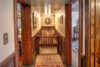 A cozy hallway with wooden paneling leads to a small landing with an ornate framed portrait hanging above the staircase. Light fixtures illuminate the space. Rooms on either side show glimpses of rustic decor and vintage furniture. A patterned rug decorates the floor.