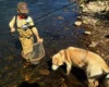 A child dressed in fishing gear, including a cap and waders, stands in a stream holding a fishing rod in one hand and a fishing net in the other. A large brown dog stands in the water beside the child, looking down into the stream. Rocks are visible in the background.