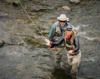 Two people standing in a river, fly fishing. The woman in the foreground is casting a fishing line while the man behind her observes. Both are wearing waders, jackets, hats, and sunglasses. The river is shallow and rocky.