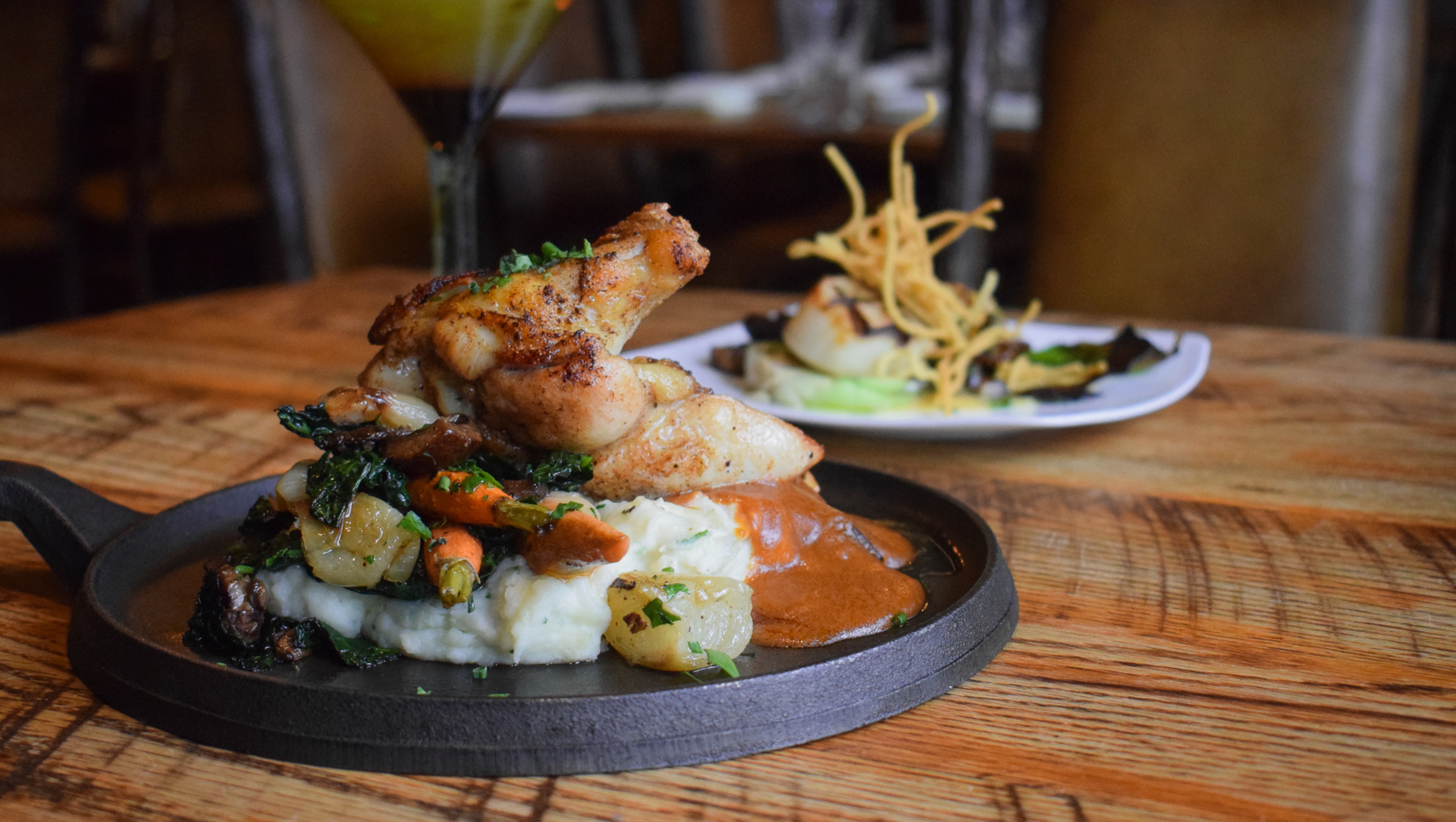 A gourmet dish of roasted chicken served on a bed of mashed potatoes and vegetables, garnished with crispy onions, presented on a black sizzling plate with a cocktail in the background.