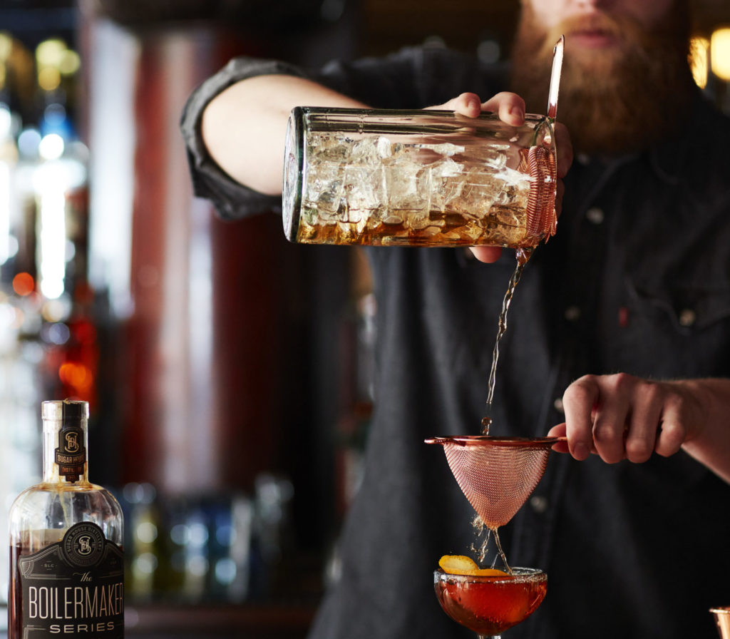 A bartender pours a dark cocktail through a strainer into a glass at a bar, with bottles visible in the background. As live music plays in Utah, he focuses intently on his task, demonstrating