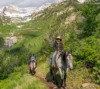 A group of people on horseback riding along a lush, mountainous trail with snow-capped peaks in the background under a clear sky.