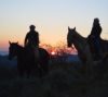 Two riders on horseback at sunset, silhouetted against a sky with hues of pink and blue, atop a mountain with a serene landscape in the background.