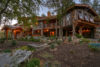 A rustic, multi-story wooden house surrounded by lush landscaping and a stone path, with warm interior lighting visible through numerous windows during twilight.