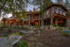 A large rustic house with wooden and stone elements during twilight. soft lights illuminate the intricate design with a lush garden in the foreground.