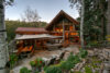 A cozy, triangular log cabin with large windows surrounded by lush trees, featuring a stone patio with outdoor furniture and soft, warm interior lighting.