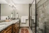 Modern bathroom featuring a glass shower, granite countertops, wooden cabinets, and a floral rug. two sinks with mirrors and towels hung neatly beside the shower.