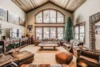 Spacious rustic-style living room with high ceilings, stone column, arch windows showing snowy landscape, leather sofas, wooden coffee table, and a large christmas tree.