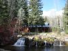 A rustic cabin beside a flowing stream with a small waterfall at Sundance Resort, surrounded by trees and brush under a clear sky.