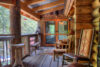 Cozy log cabin porch with rustic wooden furniture, patterned cushions, and a hanging bird feeder, surrounded by lush greenery visible through the windows and door.