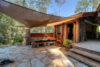 Rustic outdoor dining area with a stone table, wooden chairs, and a fabric canopy, adjacent to a wooden cabin surrounded by lush green trees.