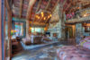 A rustic living room features exposed wooden beams, a large stone fireplace, multiple cozy seating areas with plush sofas, and an upper balcony lined with bookshelves and art. decor includes warm lighting and natural elements.