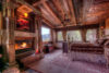 Cozy cabin-style bedroom featuring a mantled fireplace with roaring fire, ornate wooden furniture, patterned rugs, and a canopy bed draped with a luxurious fur blanket.