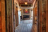 A warmly lit rustic interior hallway featuring weathered wooden doors, a worn wooden floor leading to a cozy room with a glowing fireplace and an abstract painting above it.