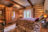 A cozy, rustic bedroom featuring log cabin walls, a large bed with a floral-patterned comforter, a wooden wardrobe, a small lamp, and a window with sheer curtains.
