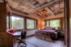 Cozy, rustic bedroom with exposed wooden beams and ceiling, large windows revealing lush greenery, a comfortable bed, desk area, and warm lighting.