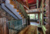 Rustic cabin interior showcasing a staircase with stone and woodwork, a vibrant rug on the floor, and a view to a greenery-framed porch through a doorway.
