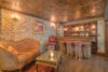 A cozy, rustic home bar with a wooden counter, red stools, and a detailed tin ceiling. a brick wall, ornate sofa, and various decorations create a warm ambiance.