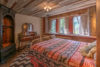 A cozy rustic bedroom with a large bed covered in a red and gray geometric pattern comforter, wooden beam ceiling, exposed log walls, two windows with a forest view, and antique furniture.