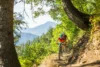 Two mountain bikers ride on a narrow trail in a lush forest with towering green mountains in the background and sunlight filtering through the trees.
