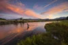 A fly fisherman in a river at sunset with dramatic pink and orange clouds above mountains, a crescent moon in the sky, and lush greenery along the banks.