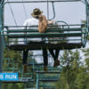 Two people on a ski lift chair, viewed from the back, ascending with trees in the background. One wears a hat, and both dangle their legs casually while enjoying summer activities in Utah.