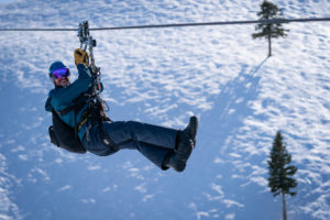 A man in winter gear zip-lines across the snow covered mountains.