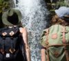 Two individuals with backpacks gaze at a waterfall while hiking in Utah. One sports a green hat and the other a silver cap, surrounded by nature.