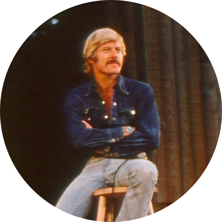 A 1970's Robert Redford sits on a stool with his arms crossed.