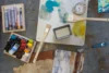 Top view of an artist's workspace with paint tubes, brushes, a palette, a mixing tray, and a paint-stained table showing vibrant strokes and splatters.