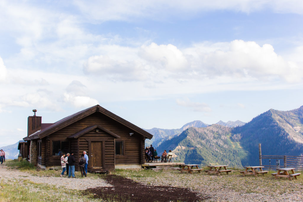 A rustic cabin with people around it located at a mountain summit, featuring expansive mountain views of surrounding peaks under a partly cloudy sky.