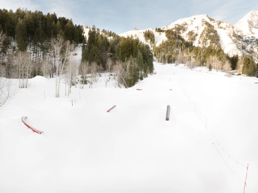 Snow-covered landscape with a ski slope featuring rails and ramps. Trees line the sides, and mountains are visible in the background under a clear blue sky.