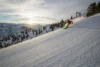 A skier in a pink jacket carving a turn on a snow-covered slope with scenic mountain views and a sunset in the background.