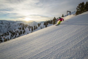 A skier in a pink jacket carving a turn on a snow-covered slope with scenic mountain views and a sunset in the background.