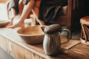 A metal pitcher with a stone lid on a wooden surface, with a brass bowl nearby and the lower legs and feet of someone seated in the background, suggesting a rustic setting.