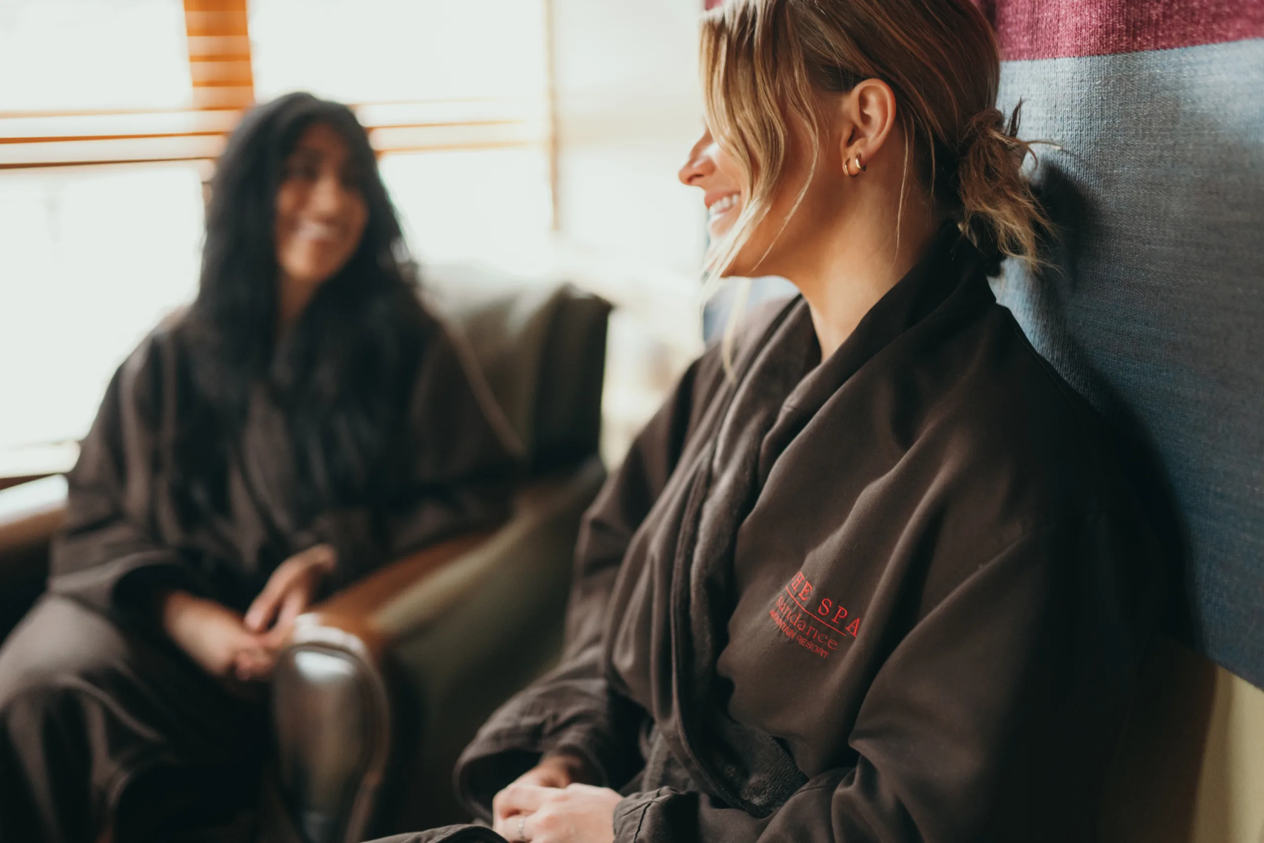 Two women smiling and relaxing in a spa setting, one blurred in the background, both wearing brown robes. comfortable and cozy ambiance with soft lighting.