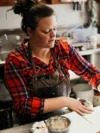 A woman in a plaid shirt and apron works attentively on a soap making project in an art studio, her hands molding clay, with tools and pots around her.