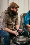 A man with a beard and a cap shaping clay on a pottery wheel in a workshop. he appears focused and engaged in his craft.