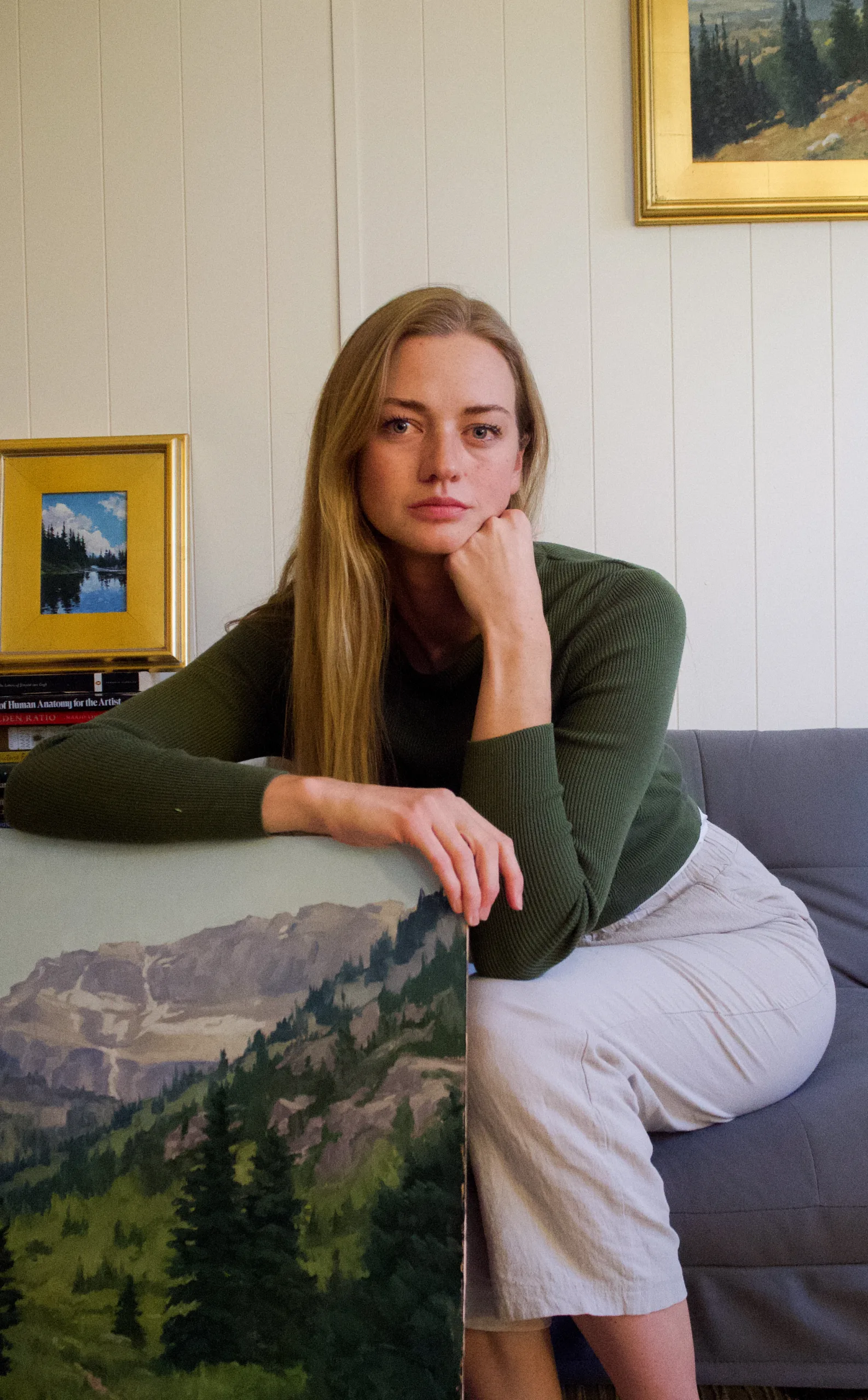 A woman with long blonde hair, wearing a dark green sweater and light pants, sits on a gray couch, resting her chin on her hand. She holds a landscape painting of mountains and trees. Framed art hangs on the wall behind her.
